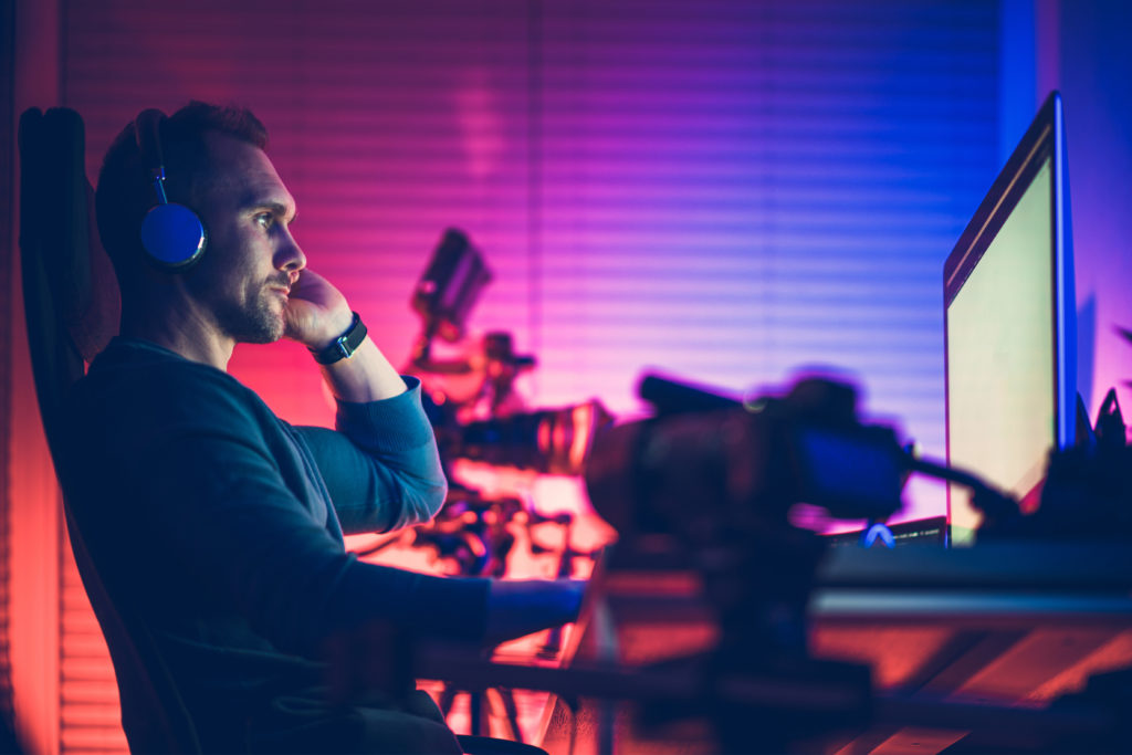 Caucasian Men in His 40s Wearing Headphones Inside His Home Office Editing Vlogs on His Desktop Computer. Cool Blue and Red Room Illumination.