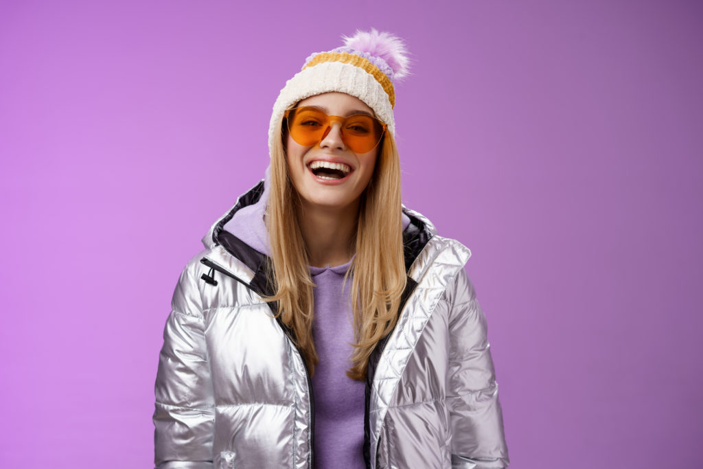 Stylish friendly charismatic blond woman in silver shiny jacket hat sunglasses ready learn snowboarding smiling laughing happily having fun snowy recreation resort, standing purple background.